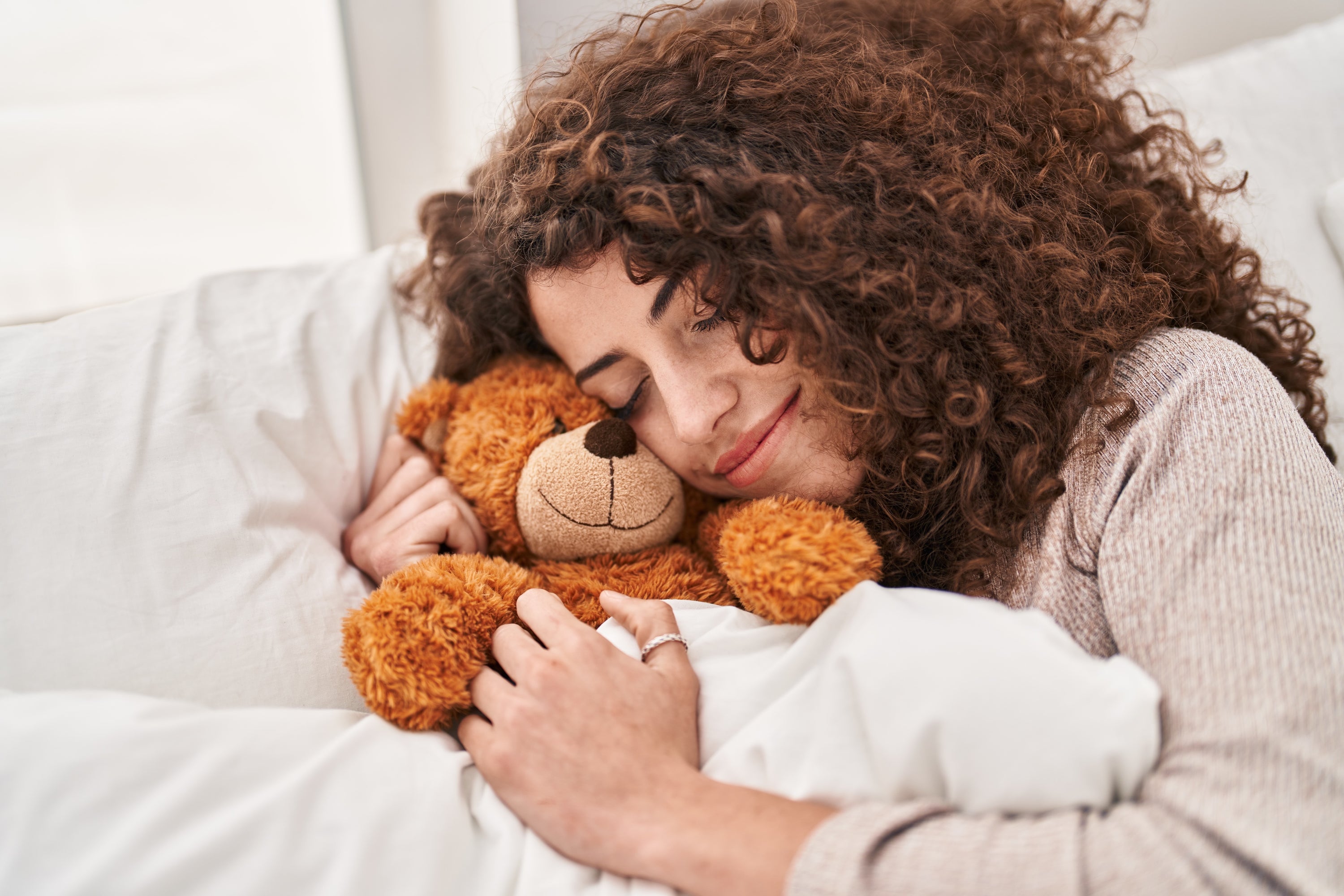 A woman laying in a white bed snuggles a teddy bear after shoulder surgery. She has curly hear and has a ring on her thumb.